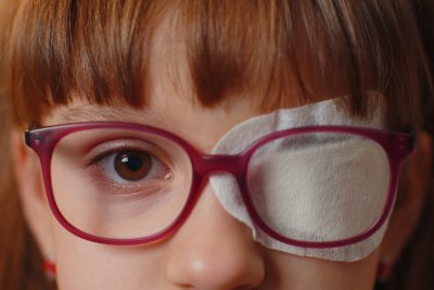 Close-up portrait of sick ill girl face looking at camera, having protective bandage plaster patch scratch on one eye. Injury, blind. Child kid in glasses. Medical eyesight vision therapy correction