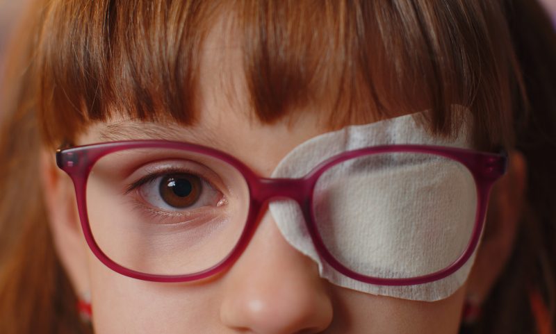 Close-up portrait of sick ill girl face looking at camera, having protective bandage plaster patch scratch on one eye. Injury, blind. Child kid in glasses. Medical eyesight vision therapy correction