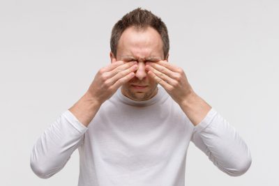 Close up of man has a problem with contact lenses, rubbing his swollen eyes due to pollen, dust allergy. Dry eye syndrome, watery, itching. Isolated on grey background, front view.