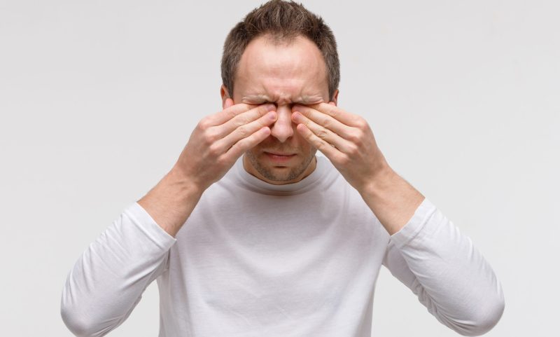 Close up of man has a problem with contact lenses, rubbing his swollen eyes due to pollen, dust allergy. Dry eye syndrome, watery, itching. Isolated on grey background, front view.
