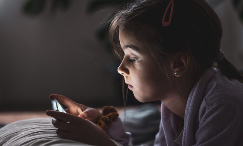 Little girl uses smartphone, lying on pillow at home, gadgets addiction concept.
